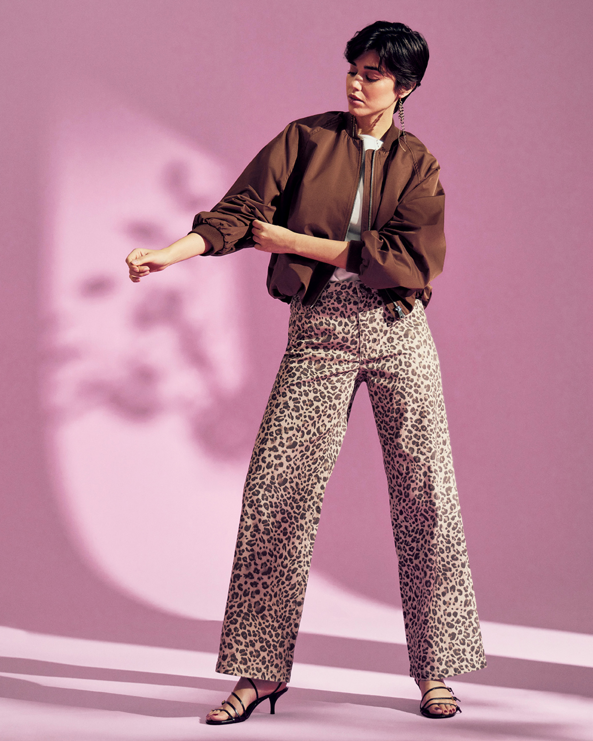 Trendiges Damen Outfit mit weiter Leo-Hose, brauner Bomberjacke und weißem Basic-Shirt vor pinkem Hintergrund.