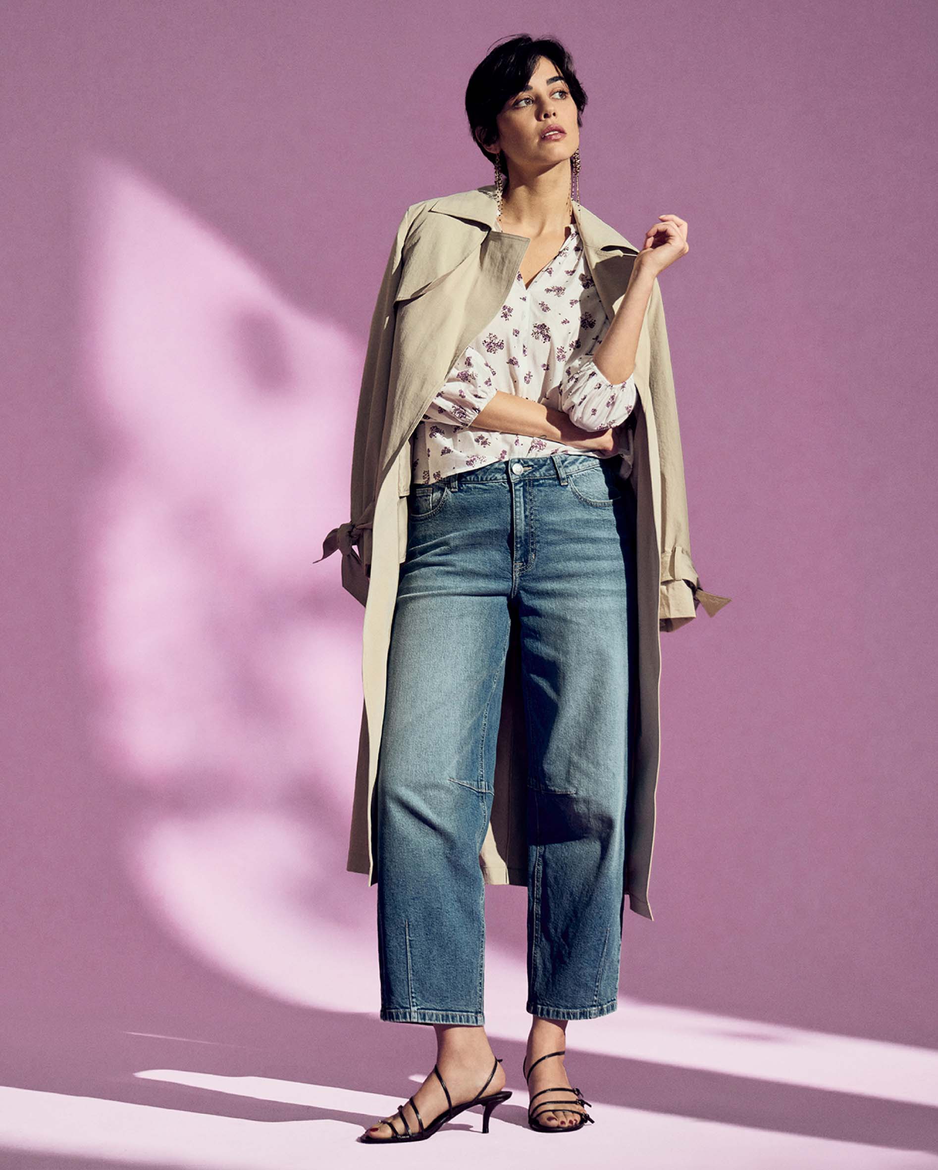 Langer Übergangsmantel mit Bindegürtel über einer Schlupfbluse mit Blumen, lässig zu Jeans gestylt, Model steht frontal.